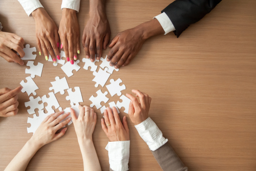 Close up of five sets of hands from employees of varying ethnicities and genders all reach for white puzzle pieces. 
