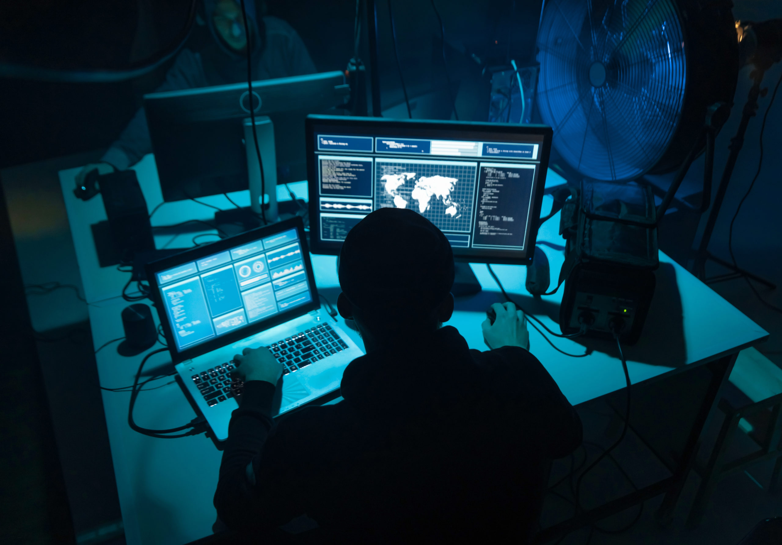 Person sits at table in dark room, using laptop computer to launch ransomware attack, looks at world map on separate monitor.