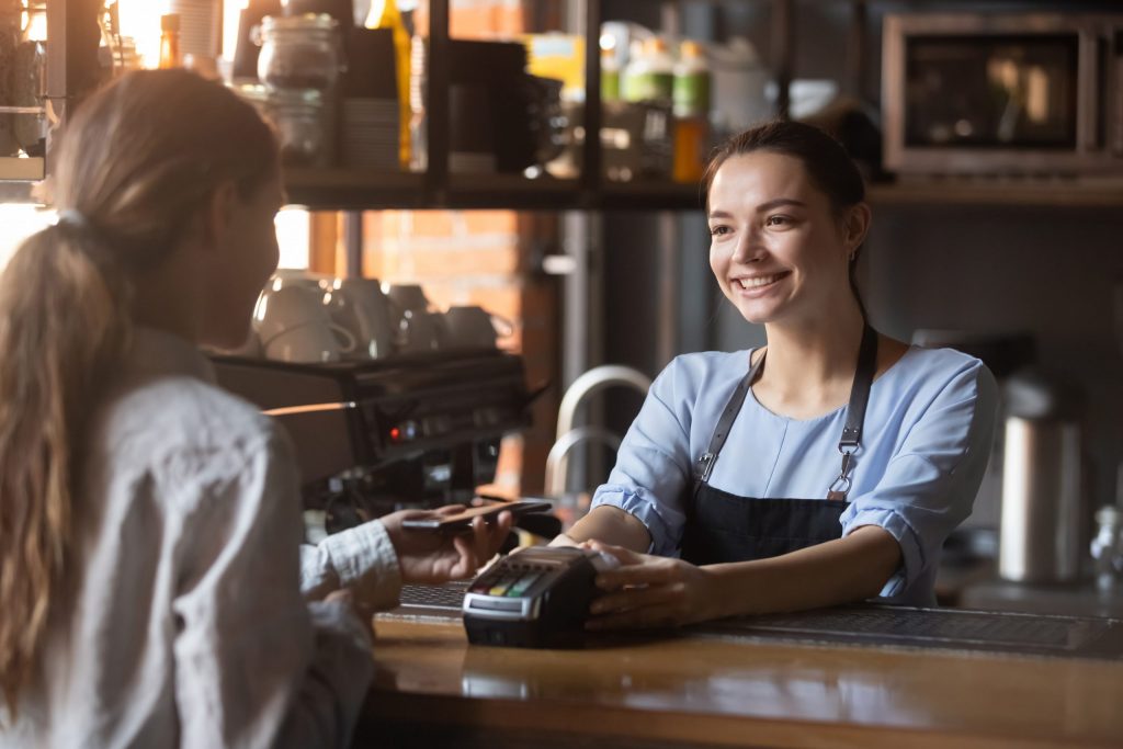 A smiling young female cashier holds a point of sale system out to a young female customer to tap and pay with her phone.