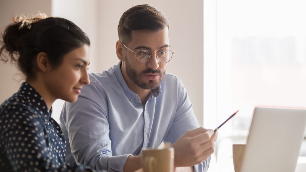 A young male and young female professional sit together at a desk and look at a white laptop to discuss Cyber Insurance.