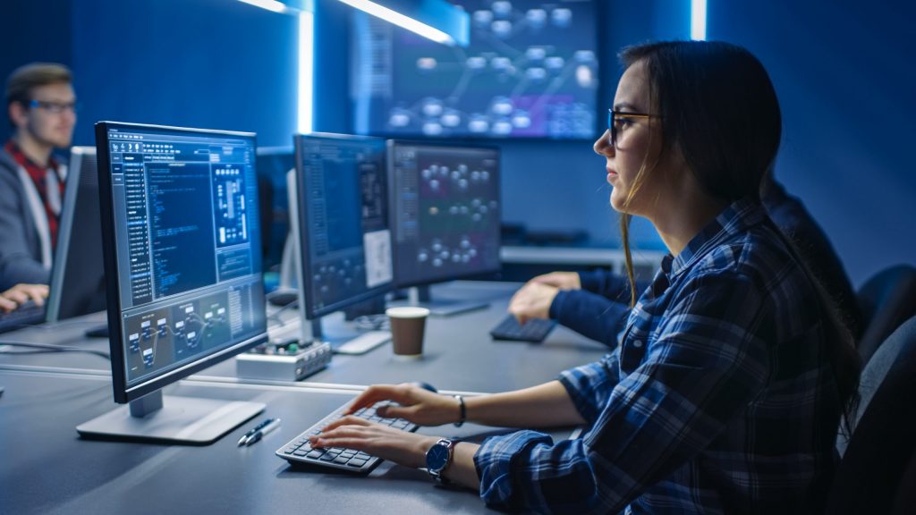 A female IT worker sits in a computer control room in front of a desktop computer.