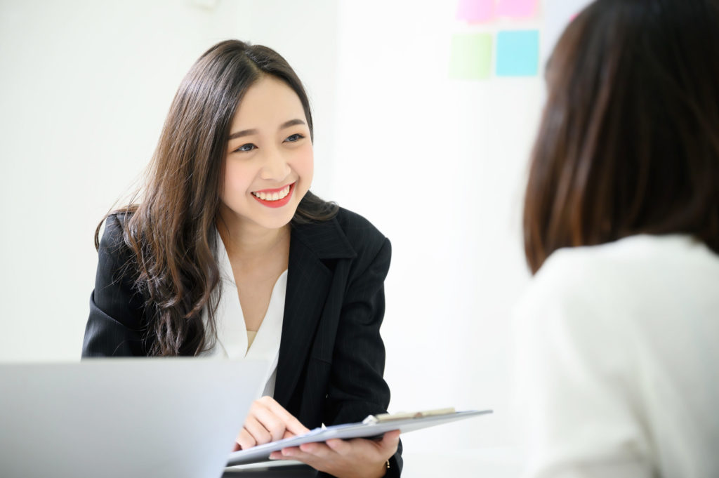 Cyber insurance broker smiles at her client from behind her laptop computer, presenting cyber insurance policy on clipboard.