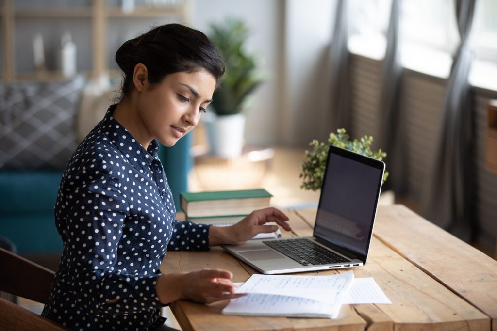 A young woman in a blue polka-dot button-down sits at home at a wooden table and works from her laptop.