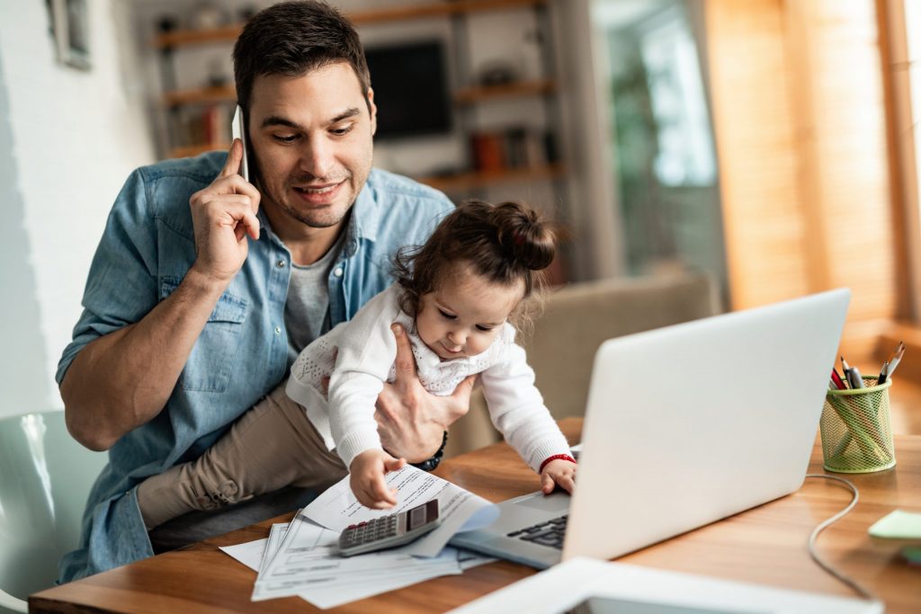 A father works from his kitchen table and holds his cell phone in one hand and his toddler daughter in the other.