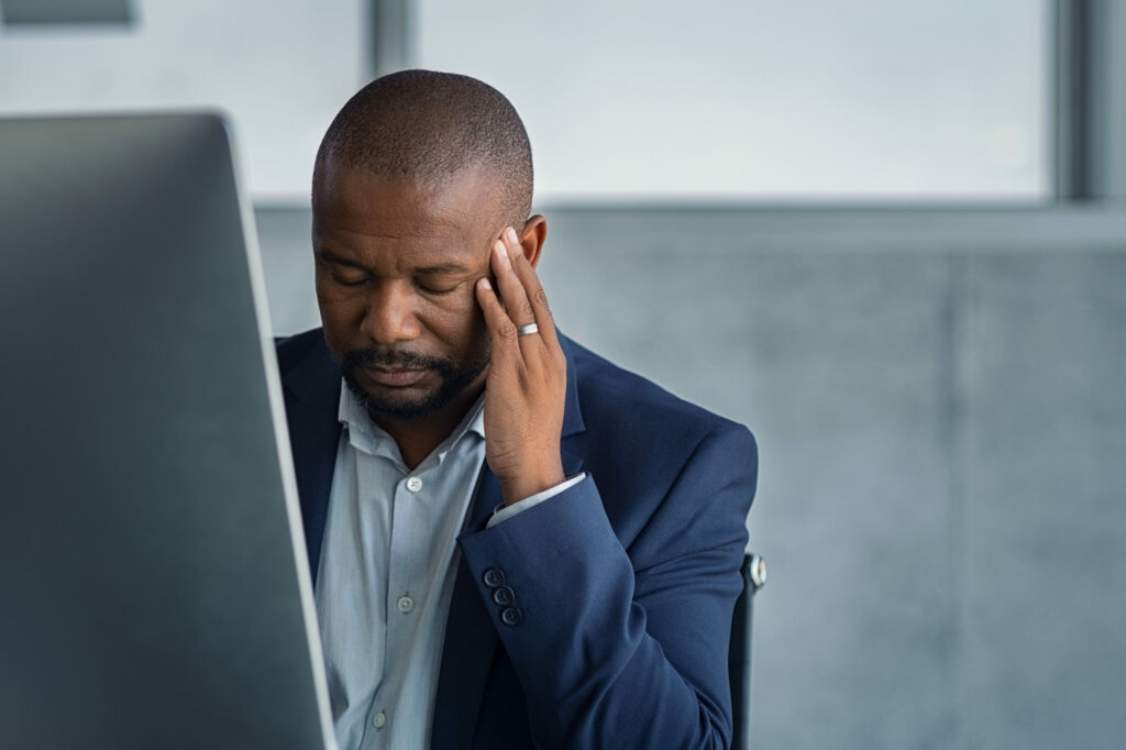  Small-business owner holds left hand to his head, looking down sadly, sitting in front of computer monitor after cyber attack.