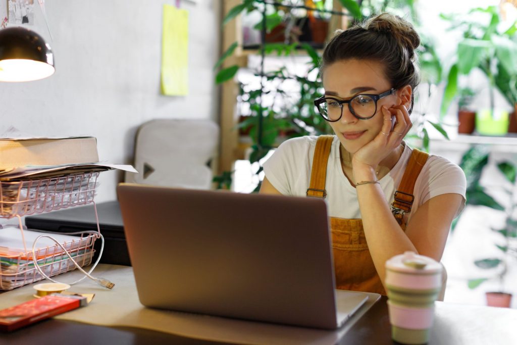 A young woman wearing orange overalls and a t-shirt works from her laptop at home.