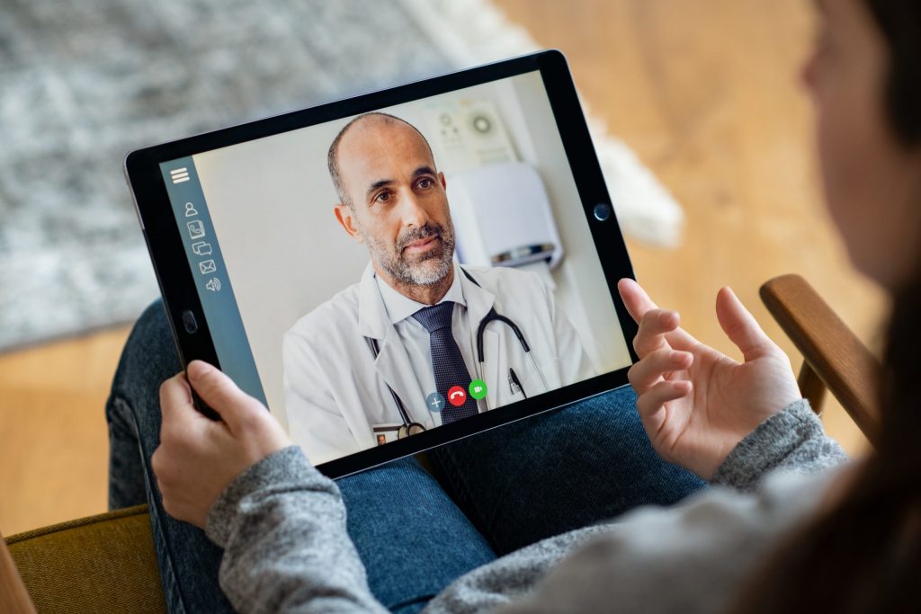 View from over the shoulder of a female patient sitting at home while video conferencing her doctor on a tablet.