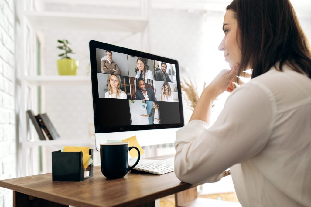 A young woman with brown hair wearing a white button-down holds a virtual meeting from her home office.