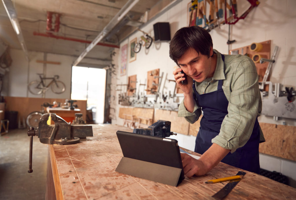Small business owner looks at laptop computer on workbench while discussing data breach insurance with his broker. 
