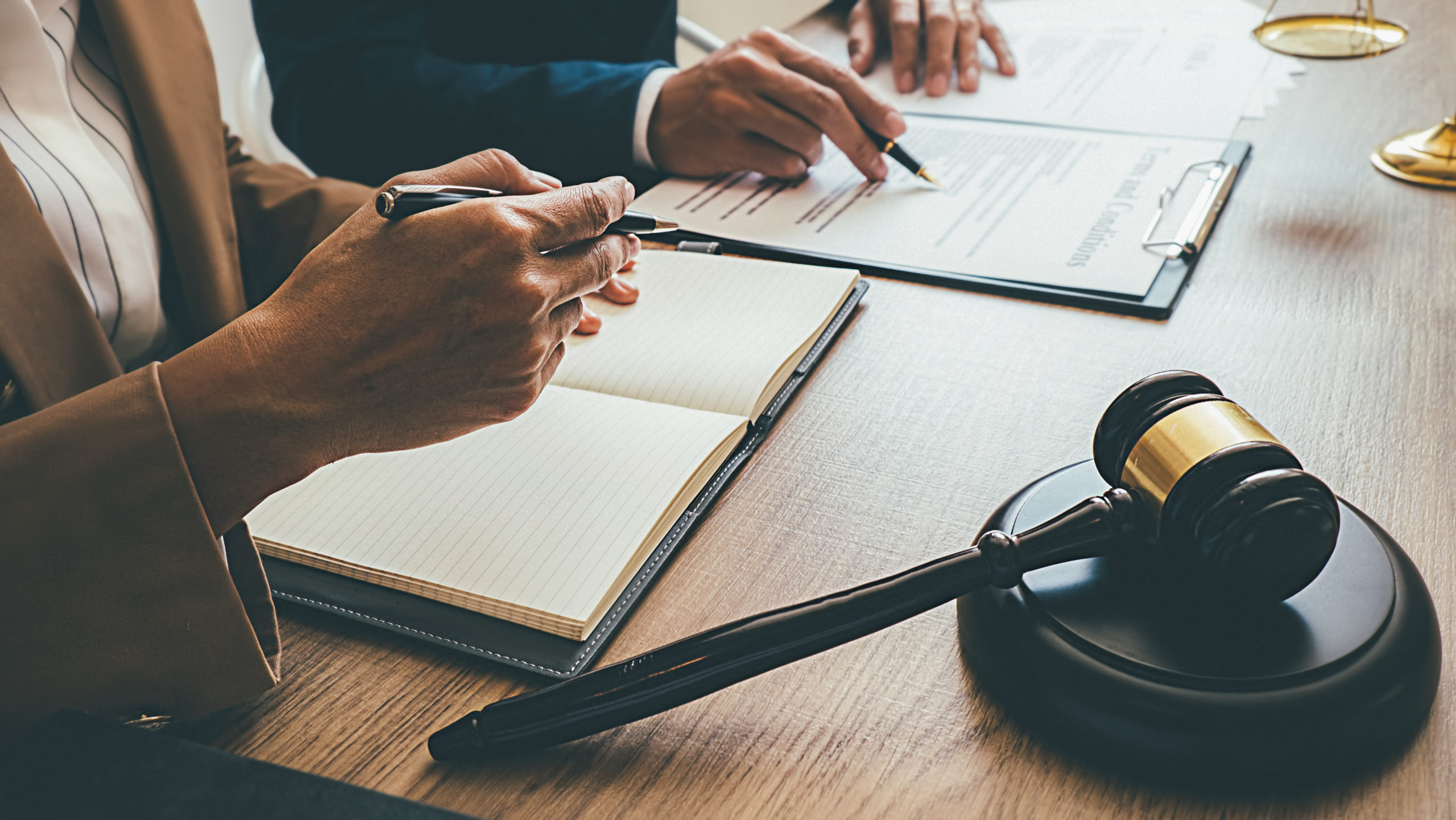 Closeup on two lawyers’ hands examining and writing notes on contracts with a wooden gavel and brass scales on table. 