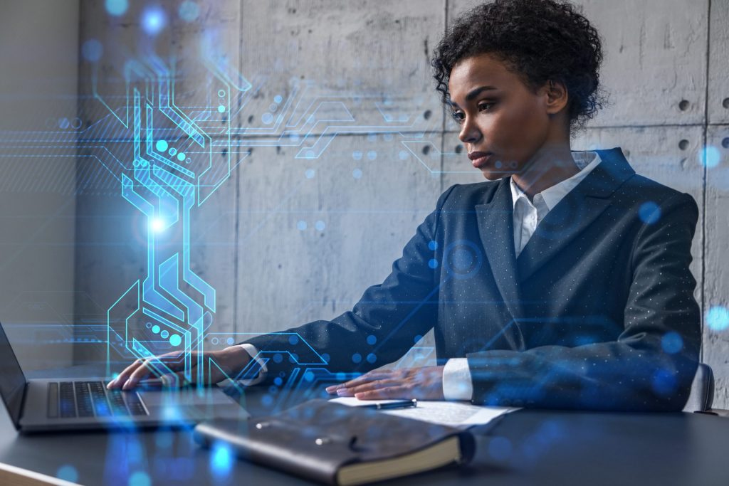 Black professional businesswoman asks what is cyber terrorism as she looks at a computer with blue tech images superimposed on it. 