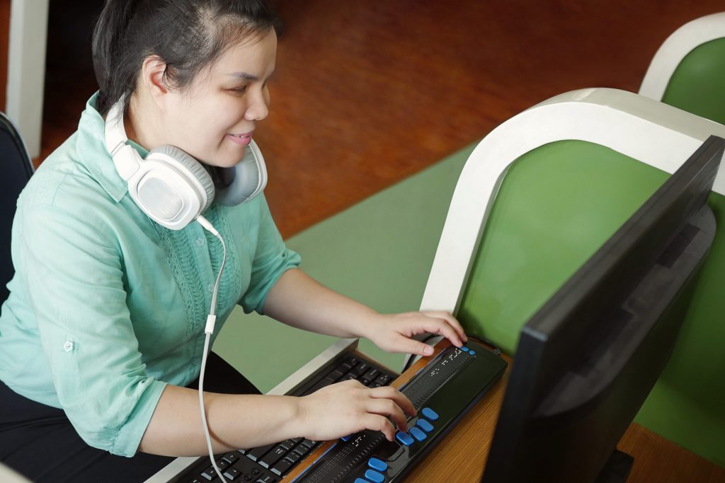 A young woman who is blind sits at a computer working on a keyboard for the visually impaired. 