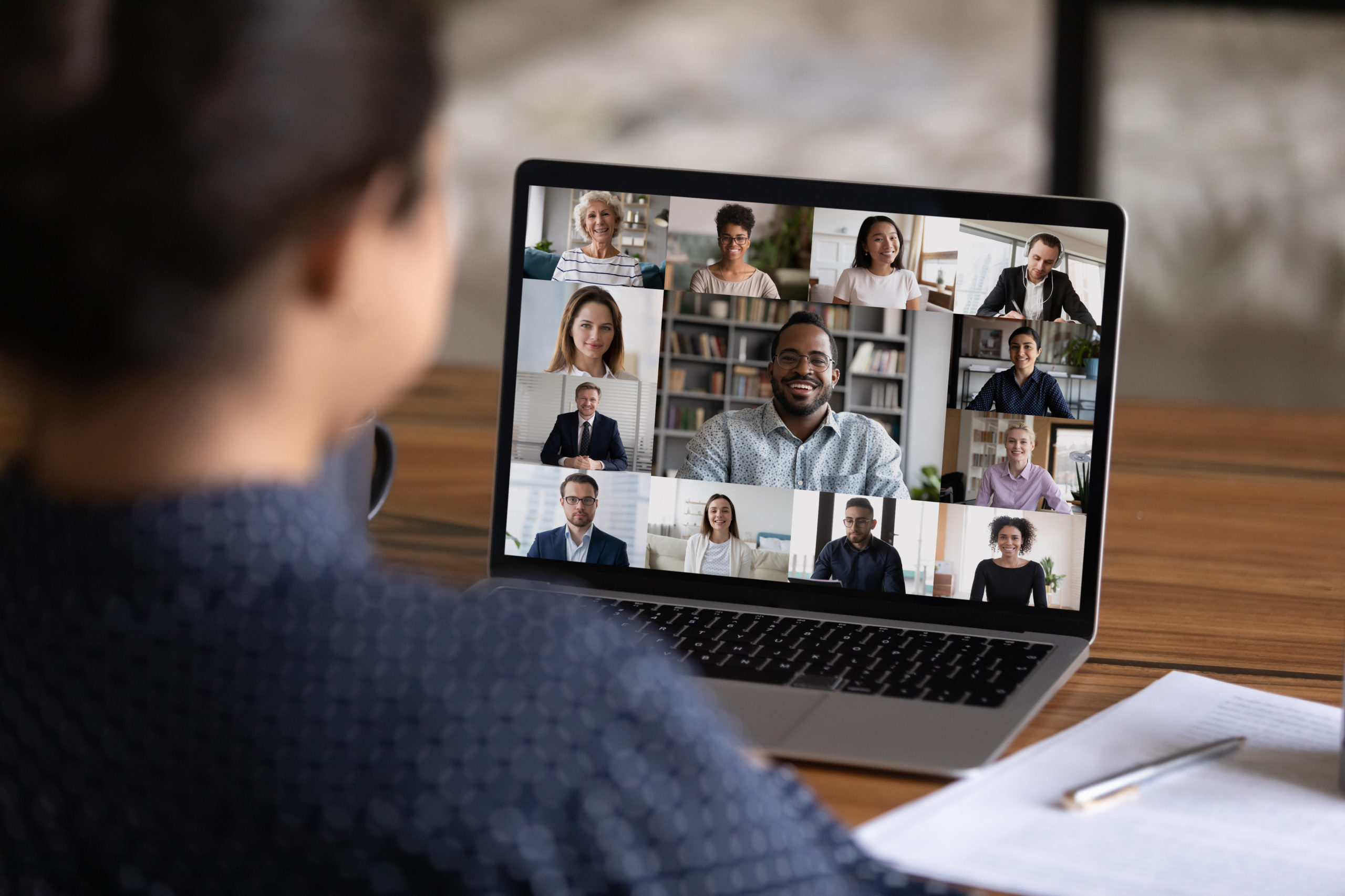 Over-the-shoulder view of woman in video conference with 13 of her colleagues, visible on screen of her laptop computer.