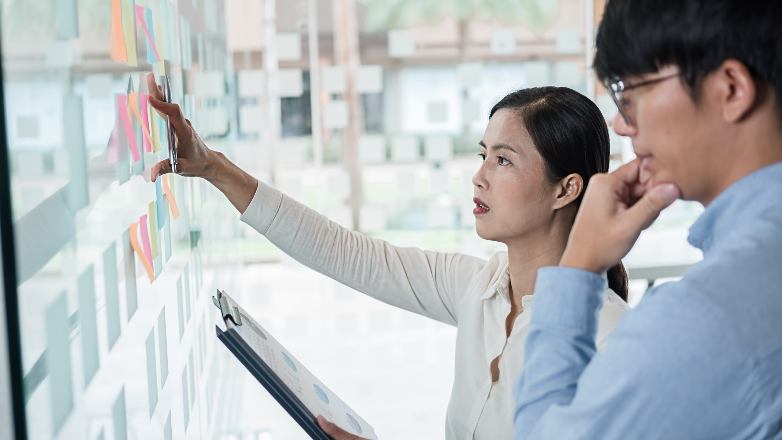 Woman and man working for a nonprofit organization stand reviewing multiple sticky notes attached to a whiteboard on office wall.