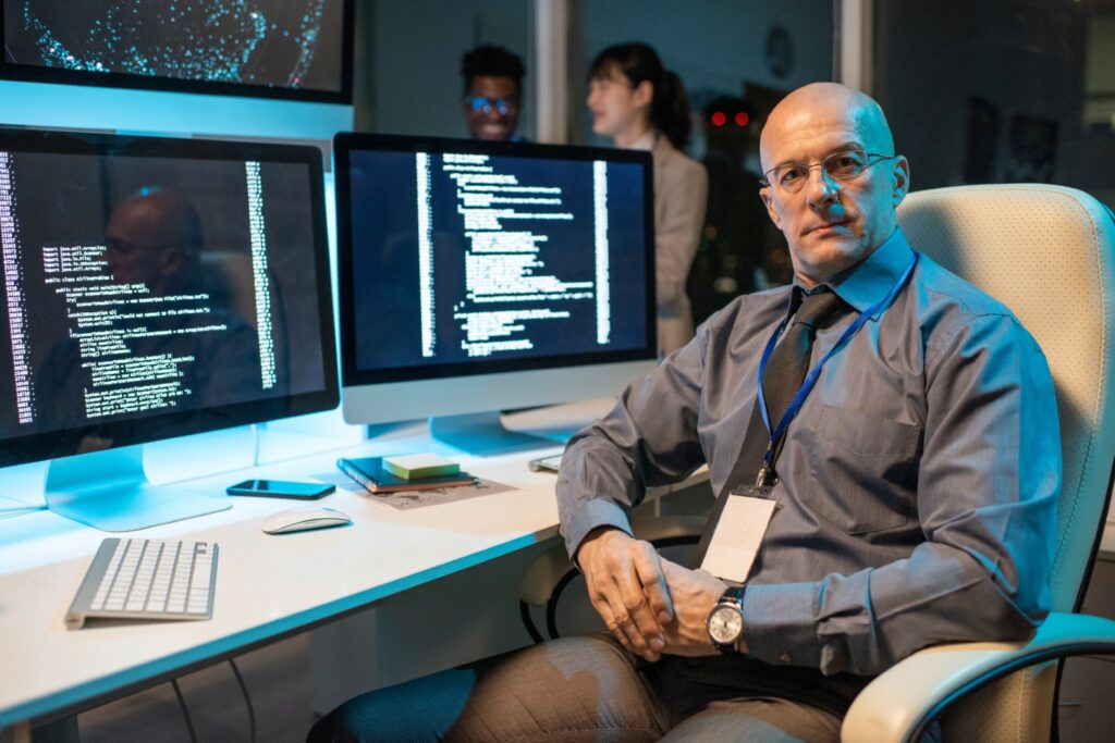 Cyber security expert sits at desk with keyboard and two computer monitors, reviewing financial services company’s risk.