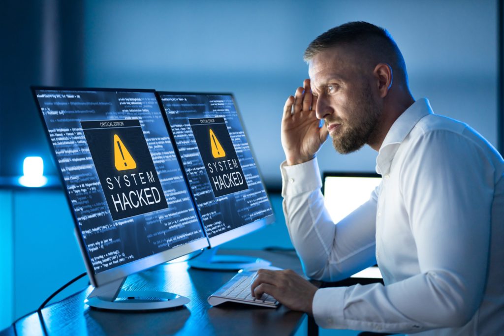 Small business owner sits at dual computer monitors showing “System Hacked” warning, illustrates need for cyber incident response.