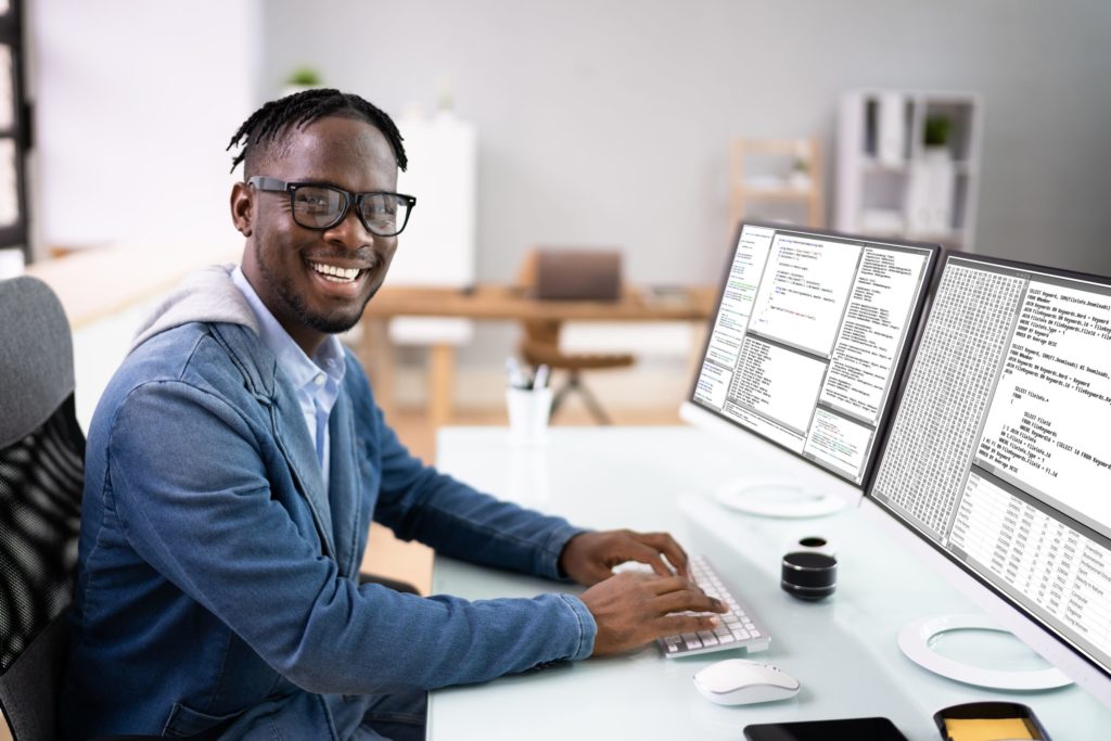 Member of computer forensics investigation team smiles at camera as he sits before two large computer monitors and keyboard.