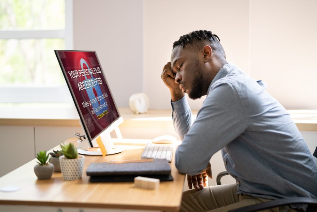 Businessman leans head on his arm and closes his eyes after reading cyber extortion message on his computer monitor.