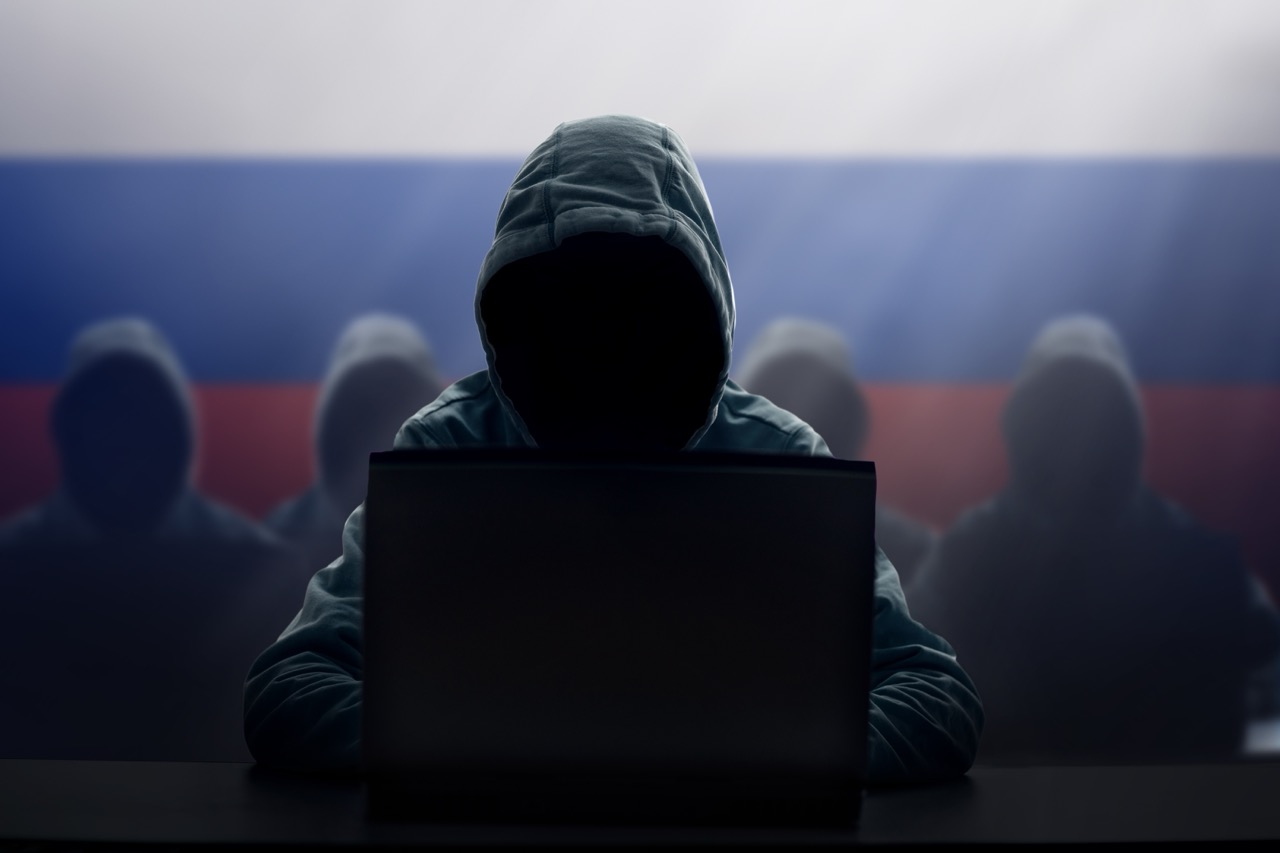 Hooded hacker at laptop with four other hooded hackers behind, all in front of Russian flag, illustrating Russian cyber attack. 