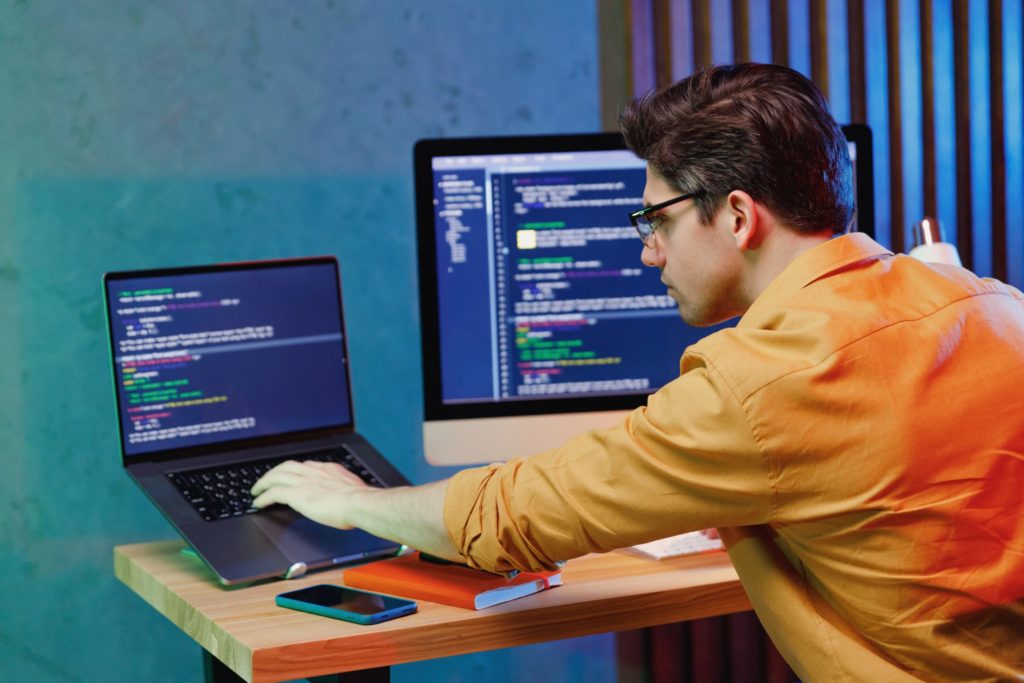 IT forensics expert reviews computer code on laptop computer, larger monitor next to laptop, after a cyber attack
