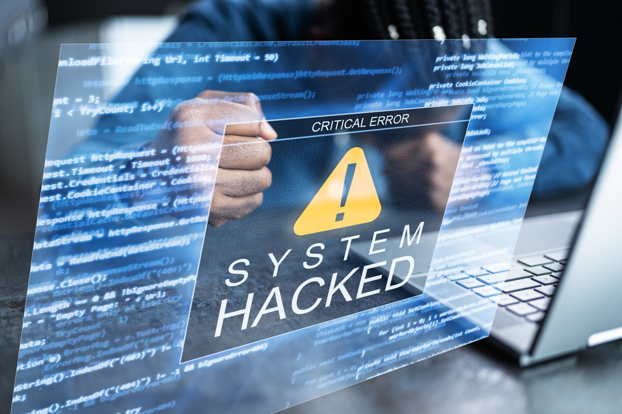 Businessman using laptop computer clenches fists in frustration, “System Hacked” warning screen overlaid on image.