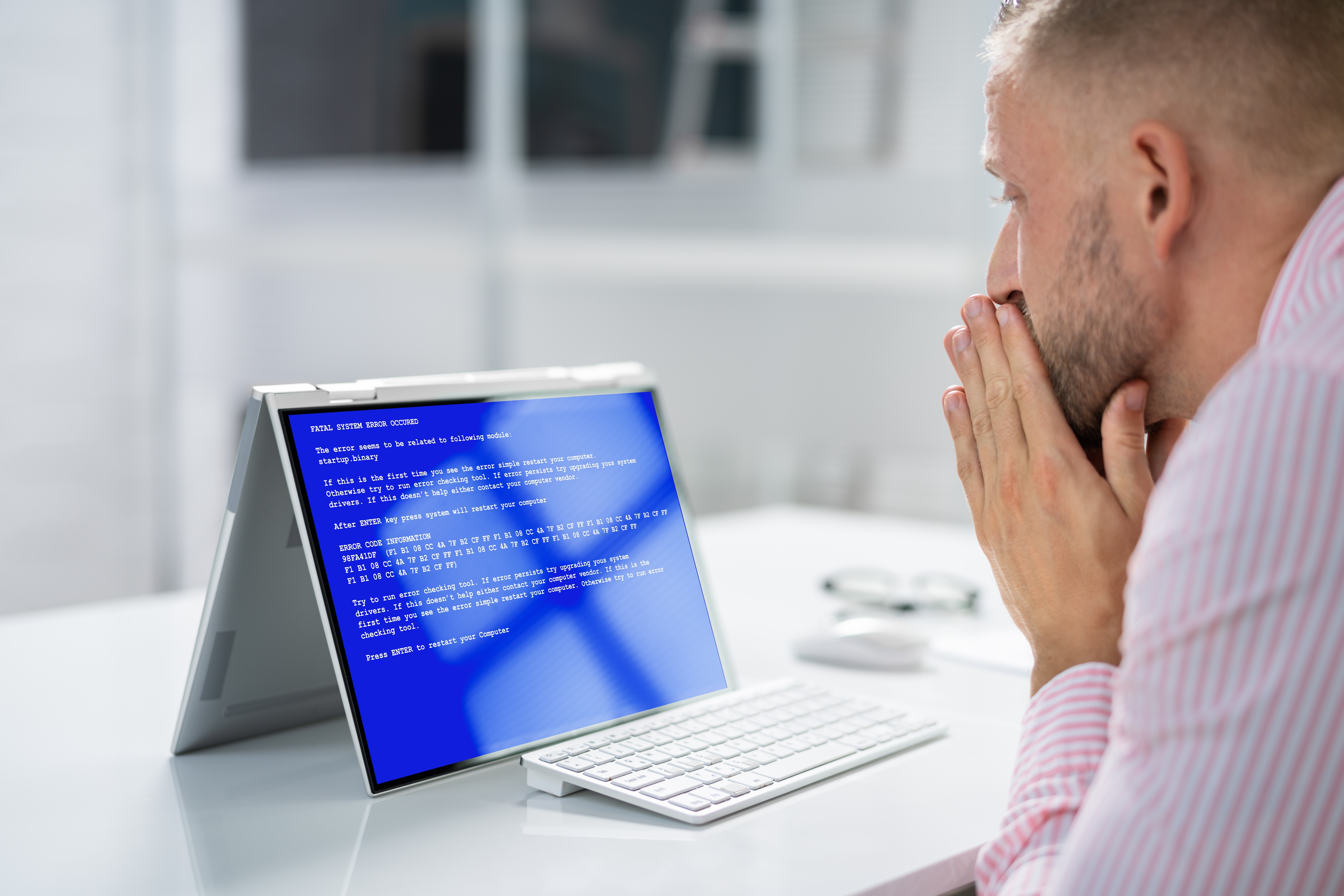 Buisnessman sits at desk, hands over his mouth in concern as his laptop monitor displays “Blue Screen of Death” error.