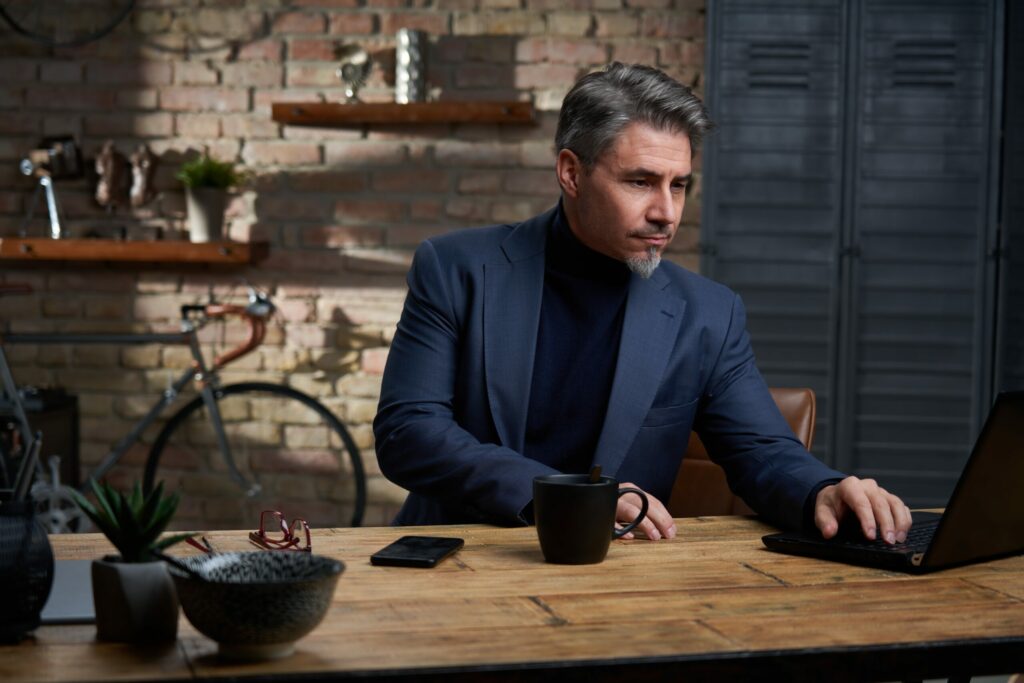 Business owner sits at rustic table using his laptop computer to review options for Cyber Insurance for small business. 