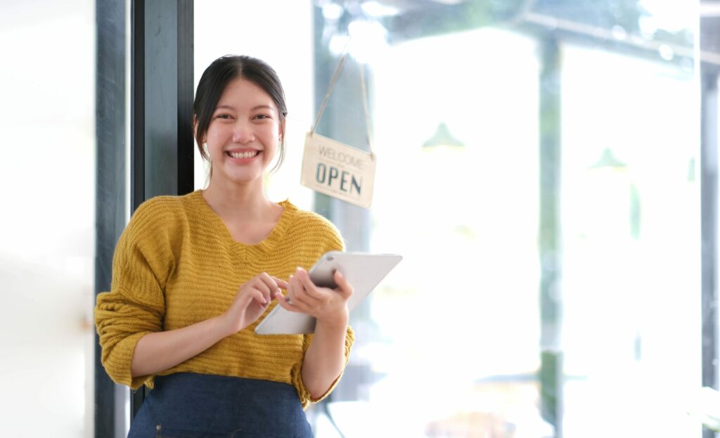 Small-business owner stands in front of her shop’s glass door, on which an “OPEN” sign hangs, using a tablet computer and smiling.