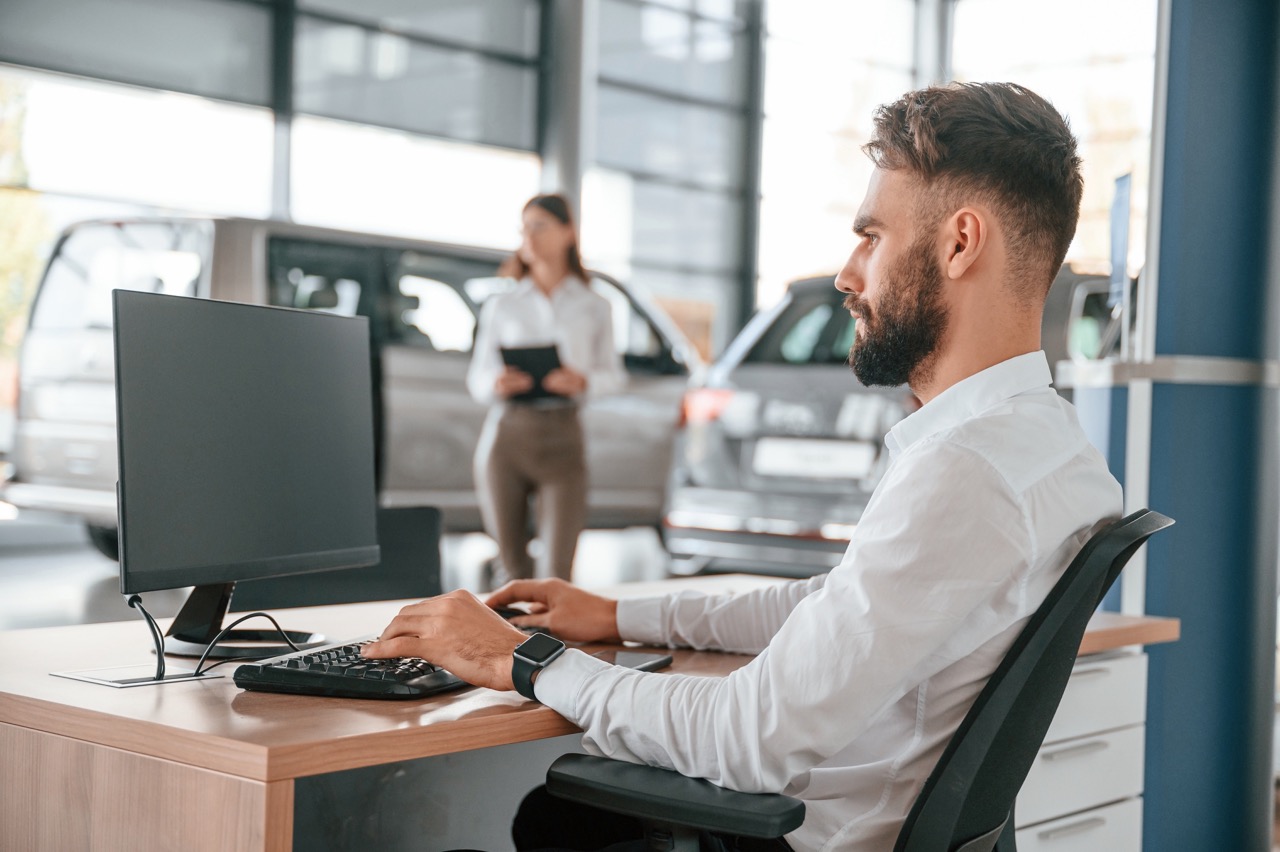 Car dealer sits at desk in showroom trying to use desktop computer during CDK Global ransomware attack. 