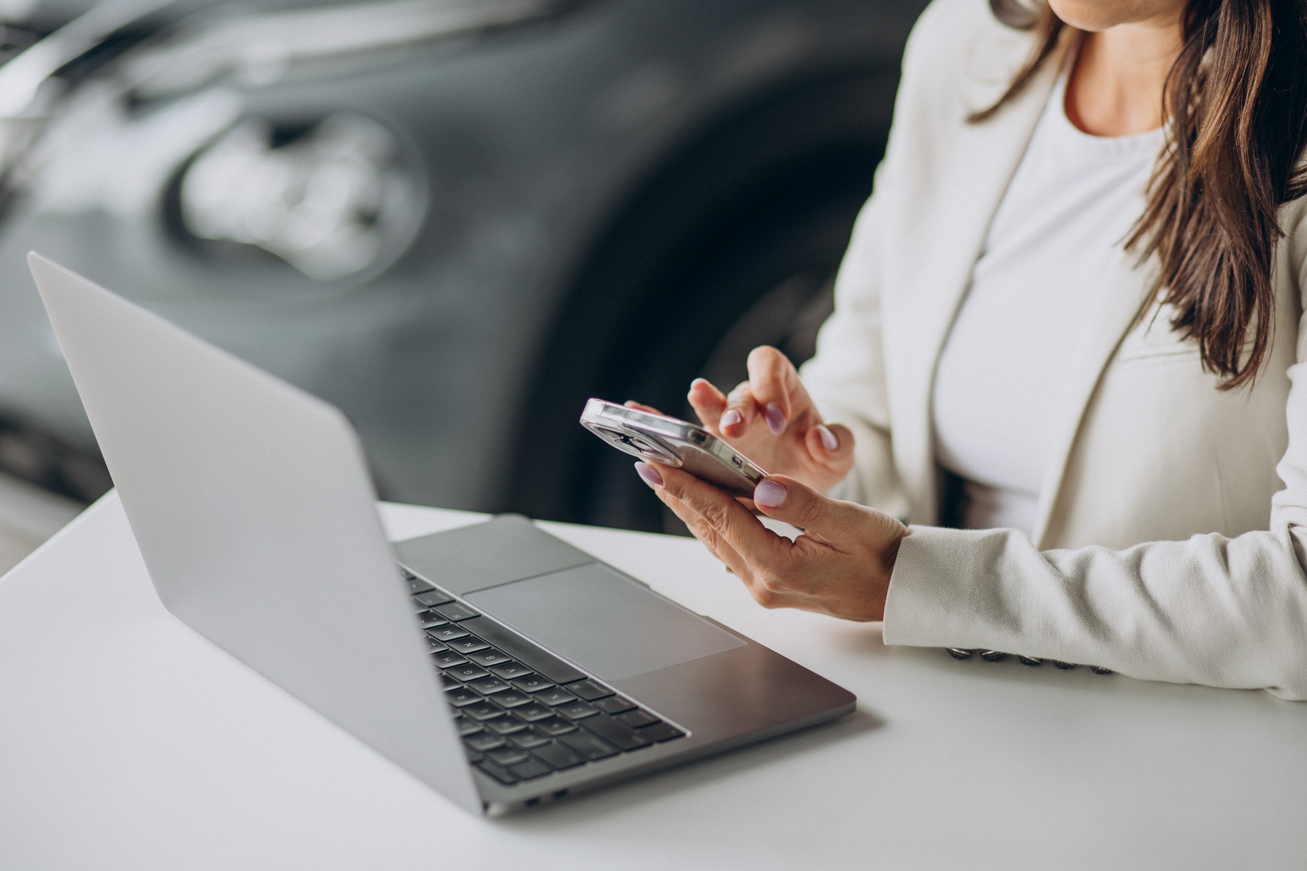 Car dealer holds her mobile phone and stands at desk with laptop computer, new car in background, during CDK ransomware attack.