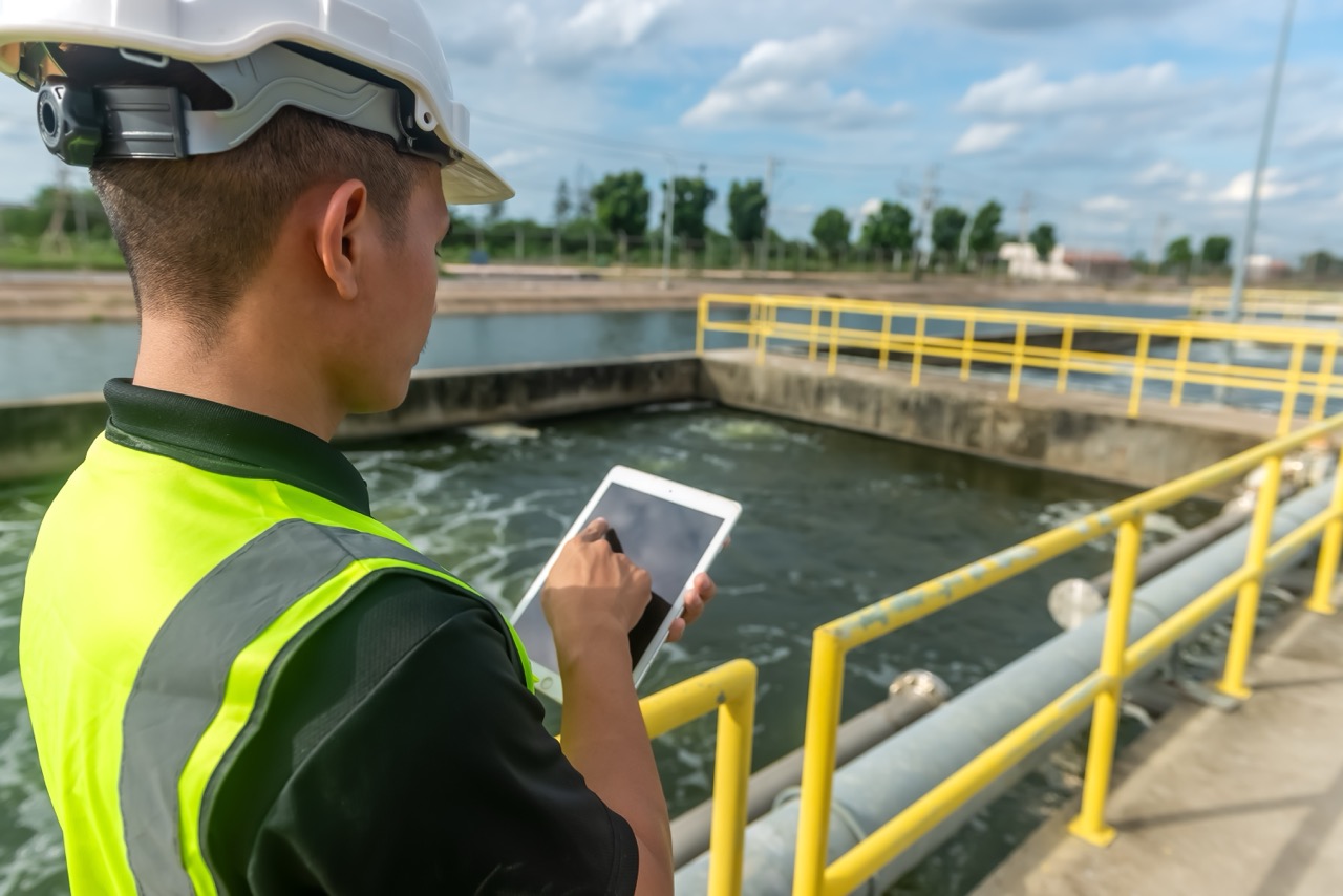 Engineer wearing hard hat and safety vest uses tablet computer to analyze water quality at public water treatment plant.