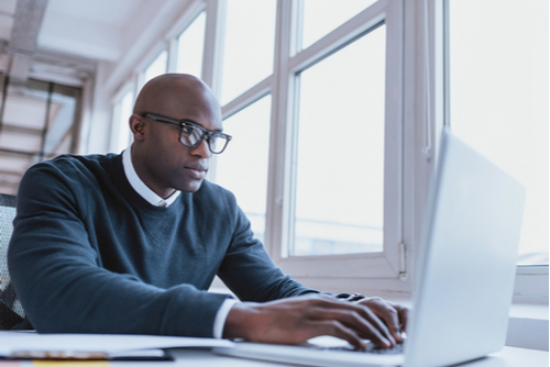 A man wearing a navy sweater and glasses types on a white laptop while at work. 