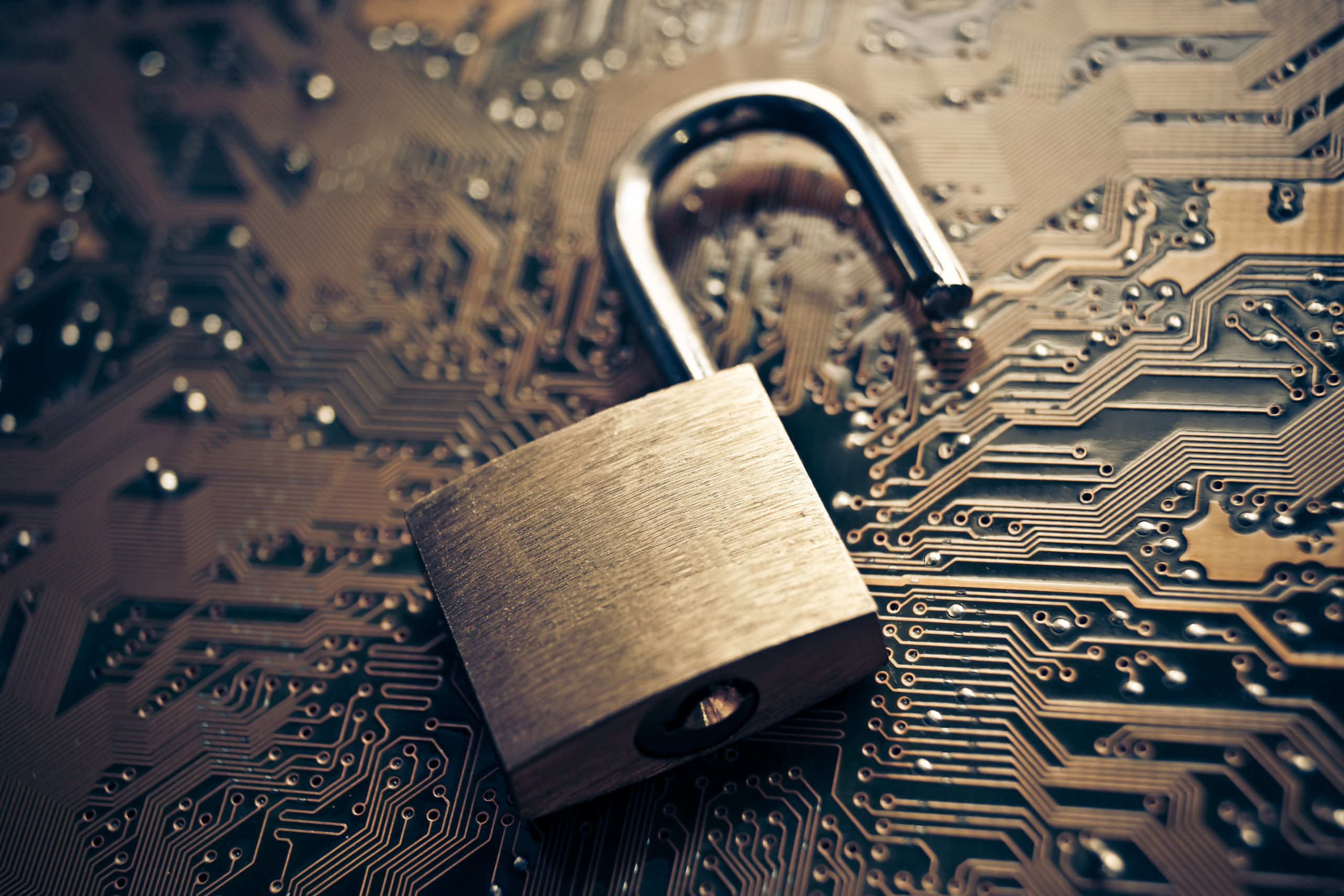 Open brass padlock on top of gold and green computer circuit board, illustrating cybersecurity concept.