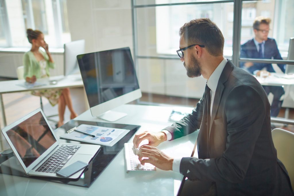 A young man in a suit sits at a white desk in an open office and types on his laptop with an extended monitor. 