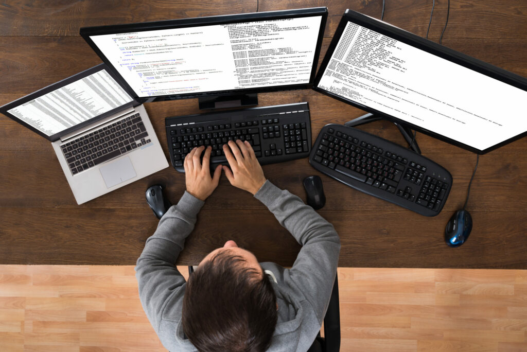  IT forensic expert sits at table using laptop computer and two large computer monitors to investigate a cyber security breach. 
