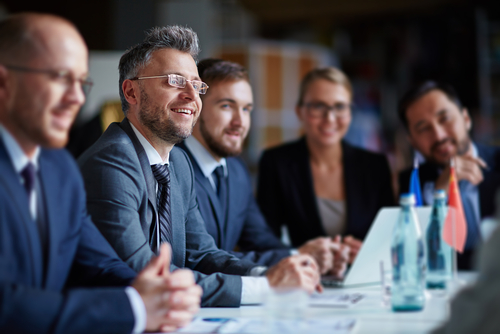 Five business colleagues in suits, four men and one woman, smile as they listen to a presentation.