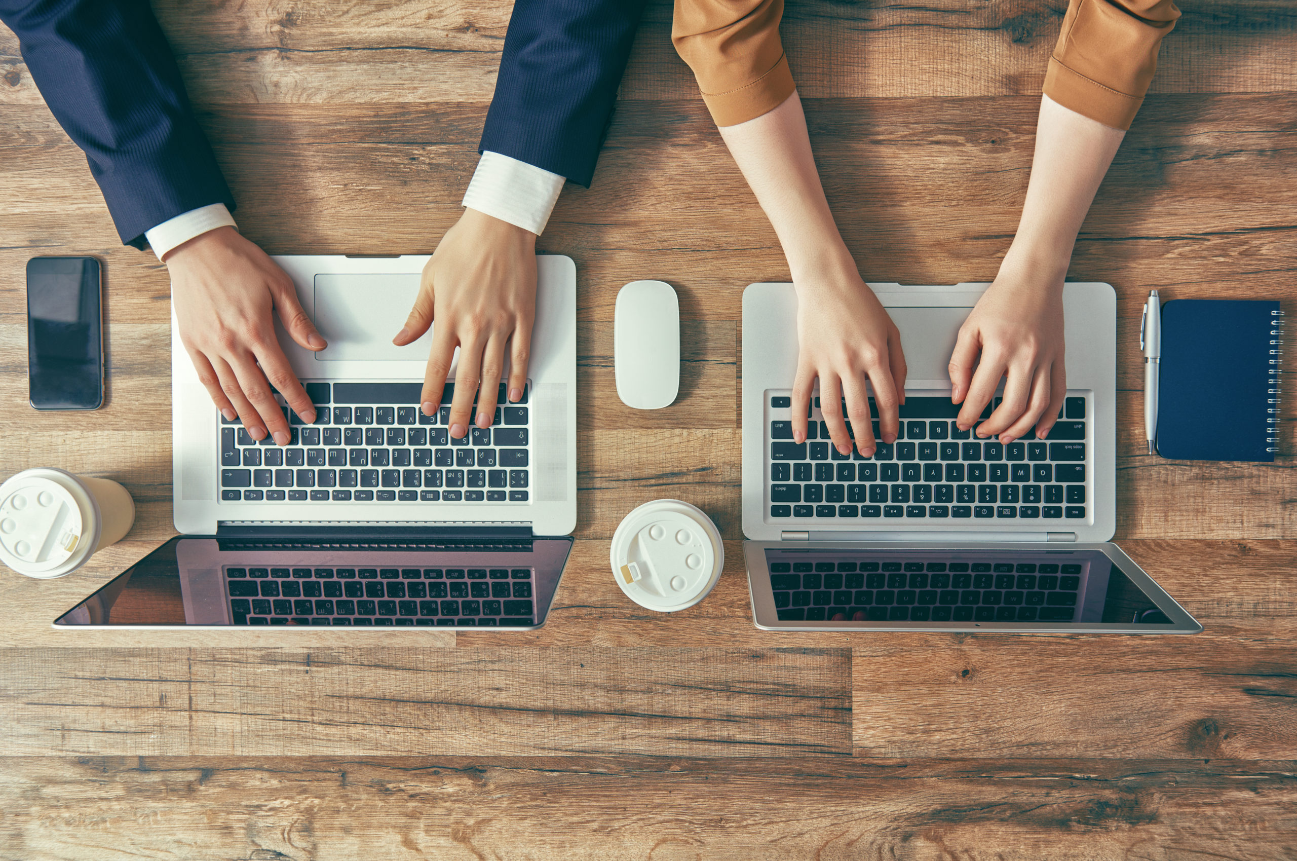 Overhead view of two pairs of businesspeople’s arms using laptop computers on wooden table, illustrating dual authorization.