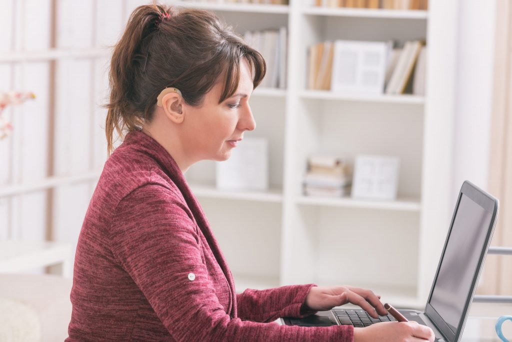 A hearing-impaired woman with a hearing aid sits at a desk and works on a laptop. 