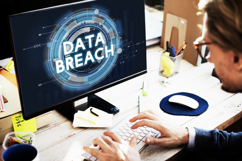Businessman sits at desk using desktop computer while monitor displays data breach warning message.