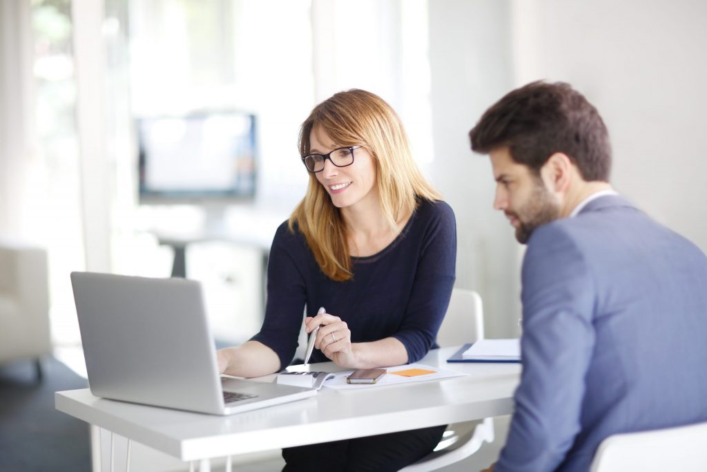 A blonde, female insurance professional shows her male client information on her white laptop. 