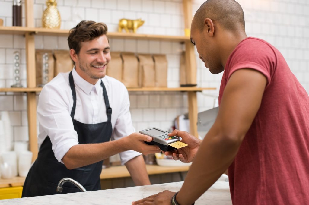 A male cashier holds a point of sale system to a male customer in a red shirt who inserts his credit card to pay. 
