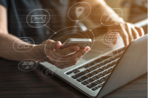 Close up of a person’s hand scrolling through a phone (which is surrounded by app graphics) while typing on a laptop. 