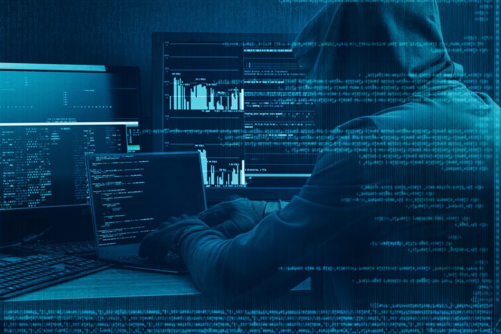 A cyber criminal in a black hoodie works at a laptop while a graphic of blue code overlays the image. 