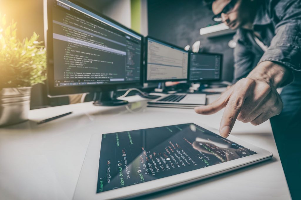 Close up of a tablet displaying code as it lies on a desk next to a desktop computer that’s also displaying code. 