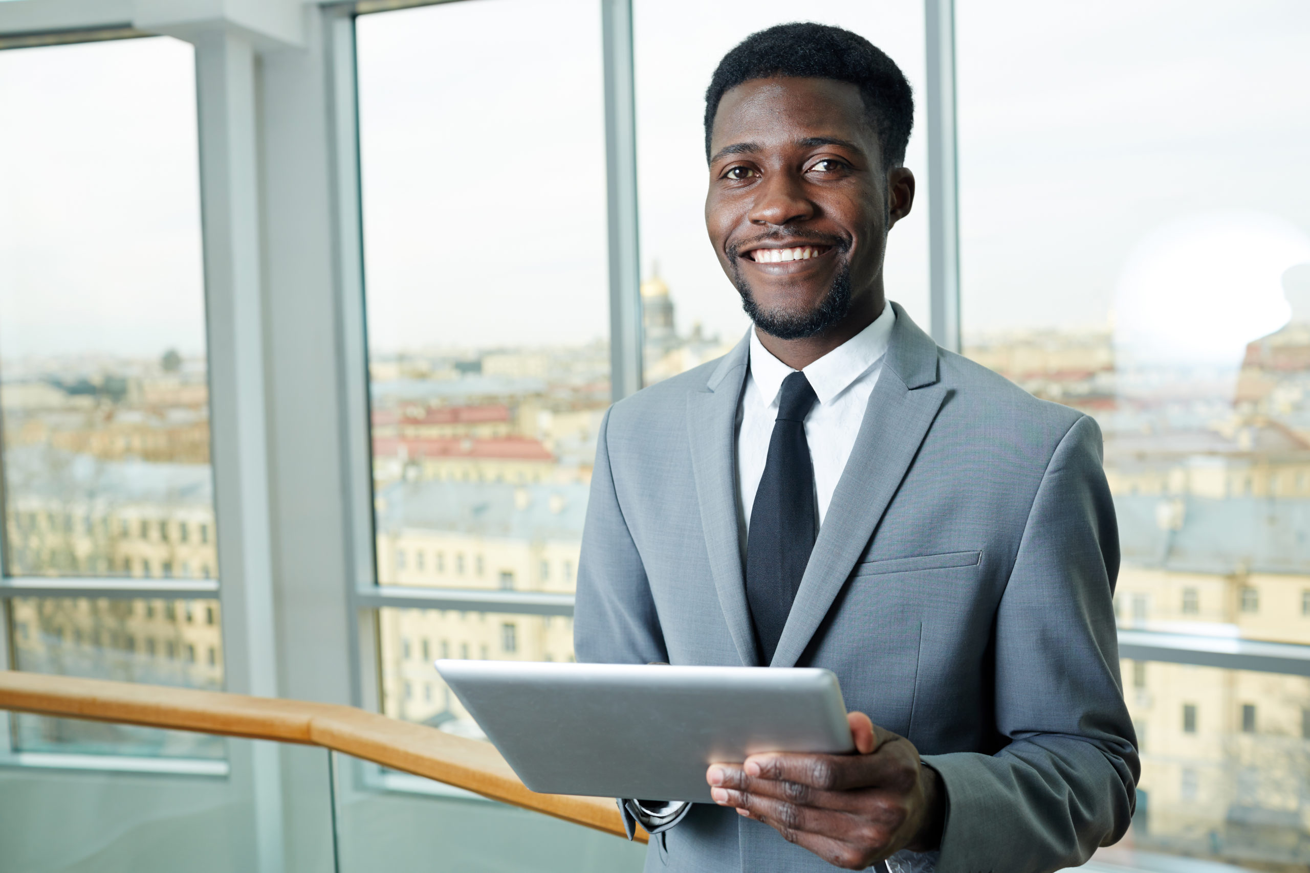 Cyber liability coverage broker smiles as he holds tablet computer and stands in front of windows in an office building. 