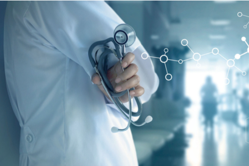 Close-up of a doctor in a white coat leaning on a hospital hallway wall with his arms crossed, holding a stethoscope. 