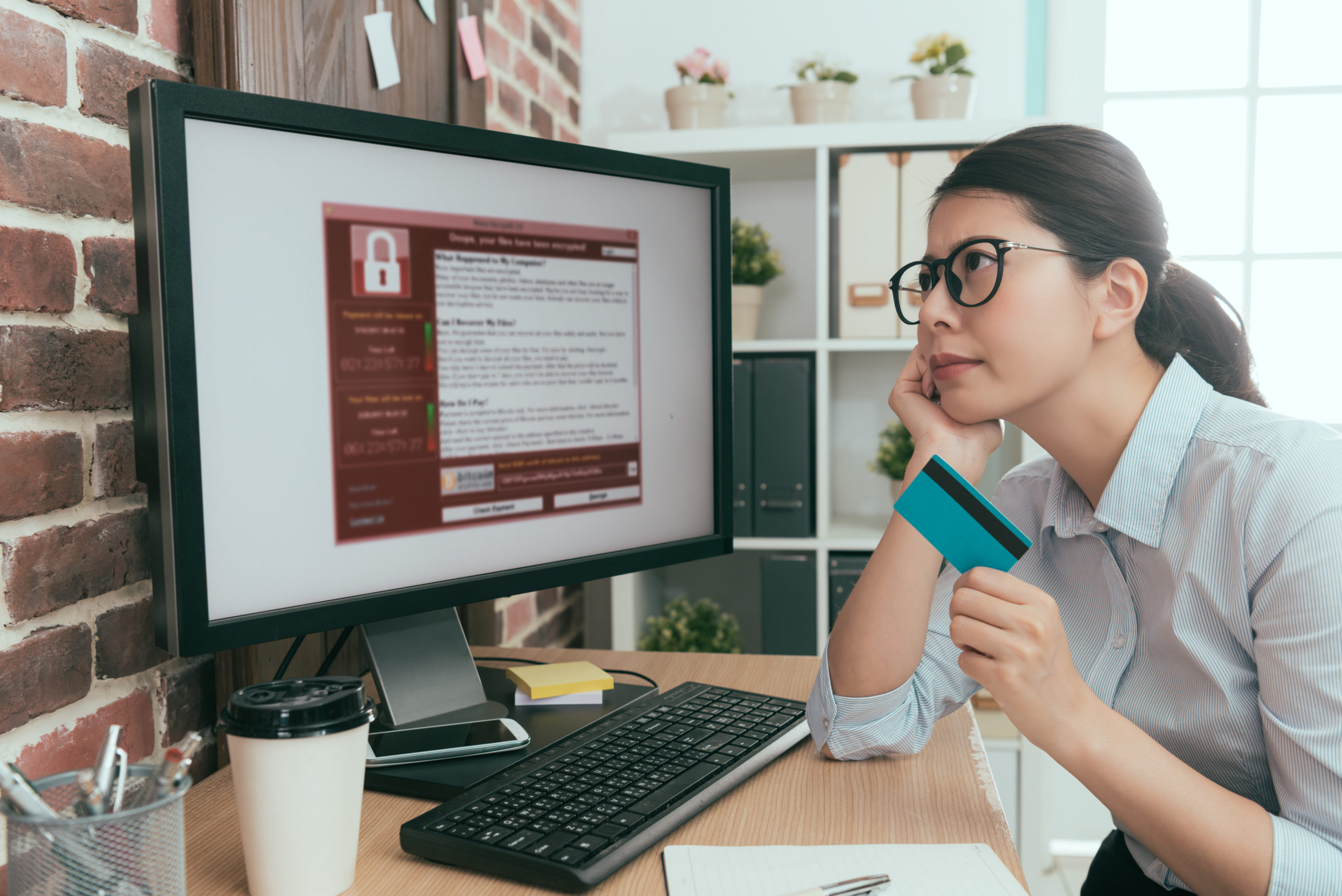  Small business owner holds her credit card and stares at her computer monitor in frustration as she pays a ransomware demand.