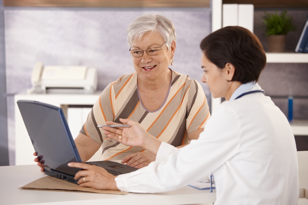 Doctor sits with senior woman in medical office, helping her understand how to log into the patient portal. 