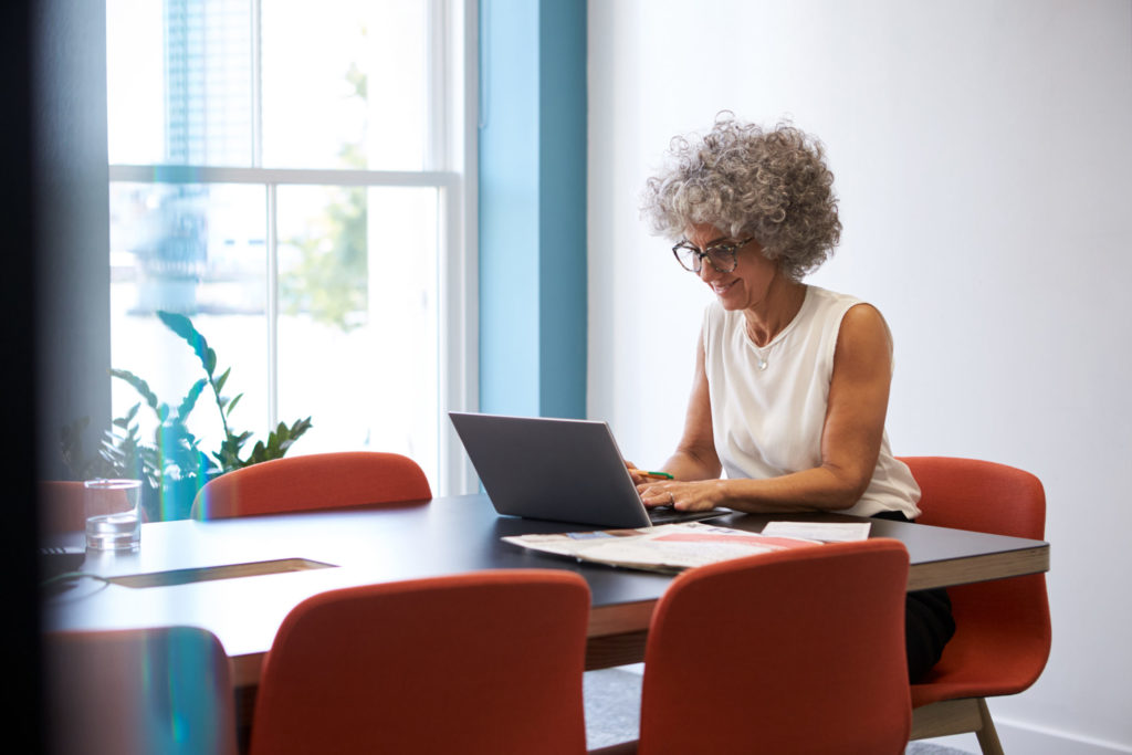 Business owner sits alone at head of conference table, smiling as she researches tech E&O insurance on laptop computer. 